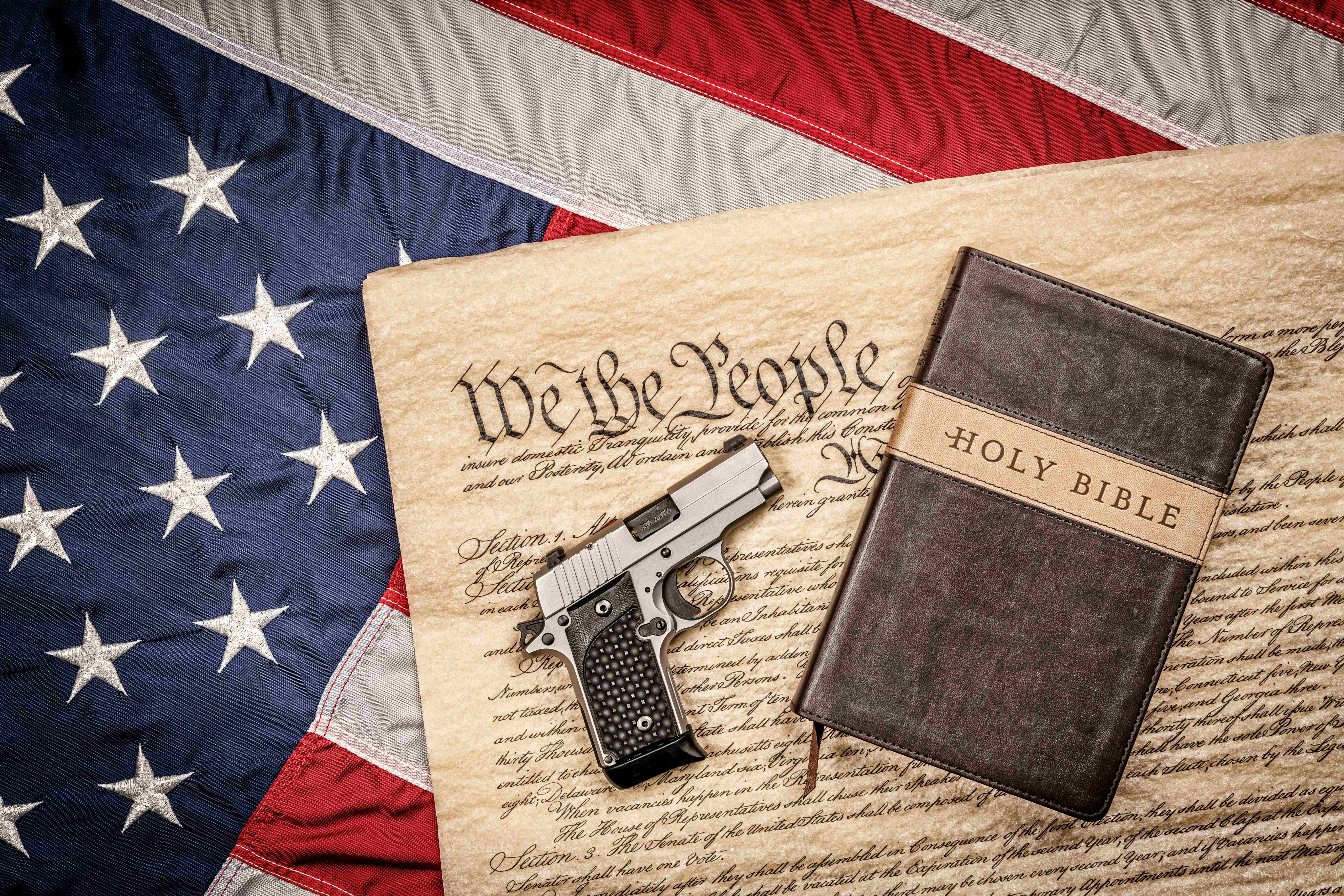 Handgun resting on the U.S. Constitution beside a Bible, all atop an American flag — symbols of faith, freedom, and constitutional rights