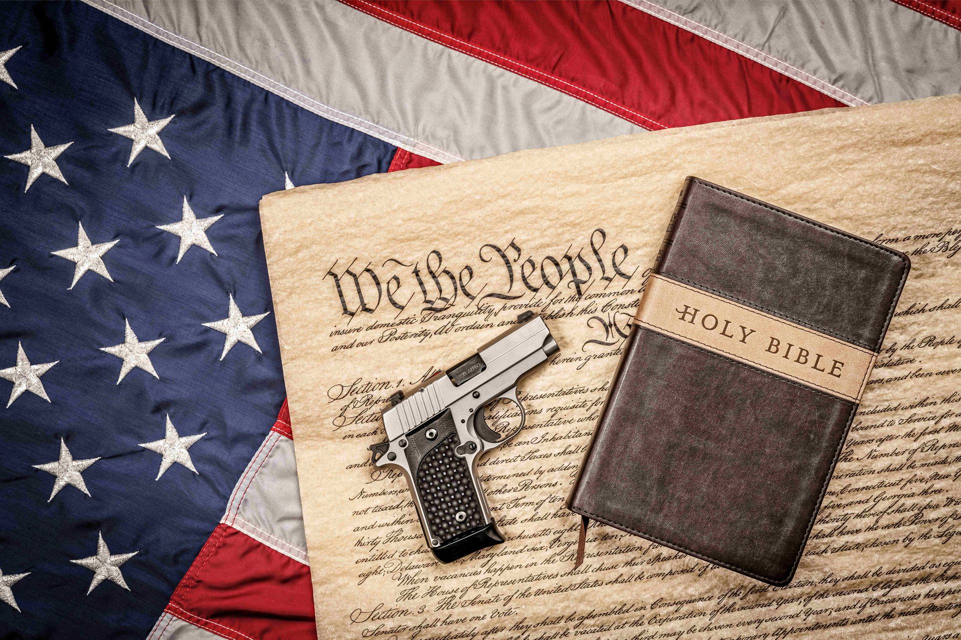 Handgun resting on the U.S. Constitution beside a Bible, all atop an American flag — symbols of faith, freedom, and constitutional rights