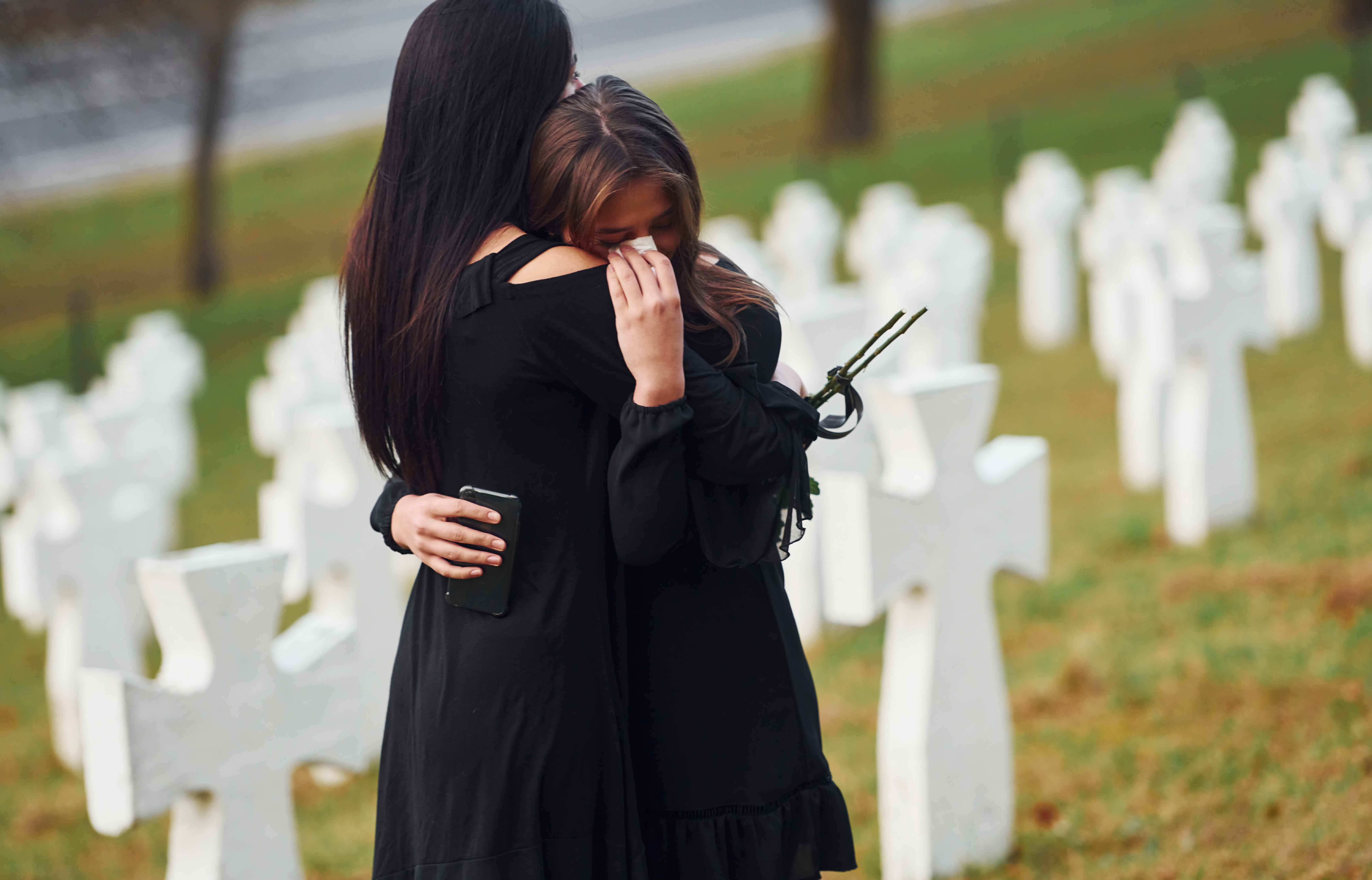 Two women in mourning attire embrace at a cemetery, comforting each other through grief