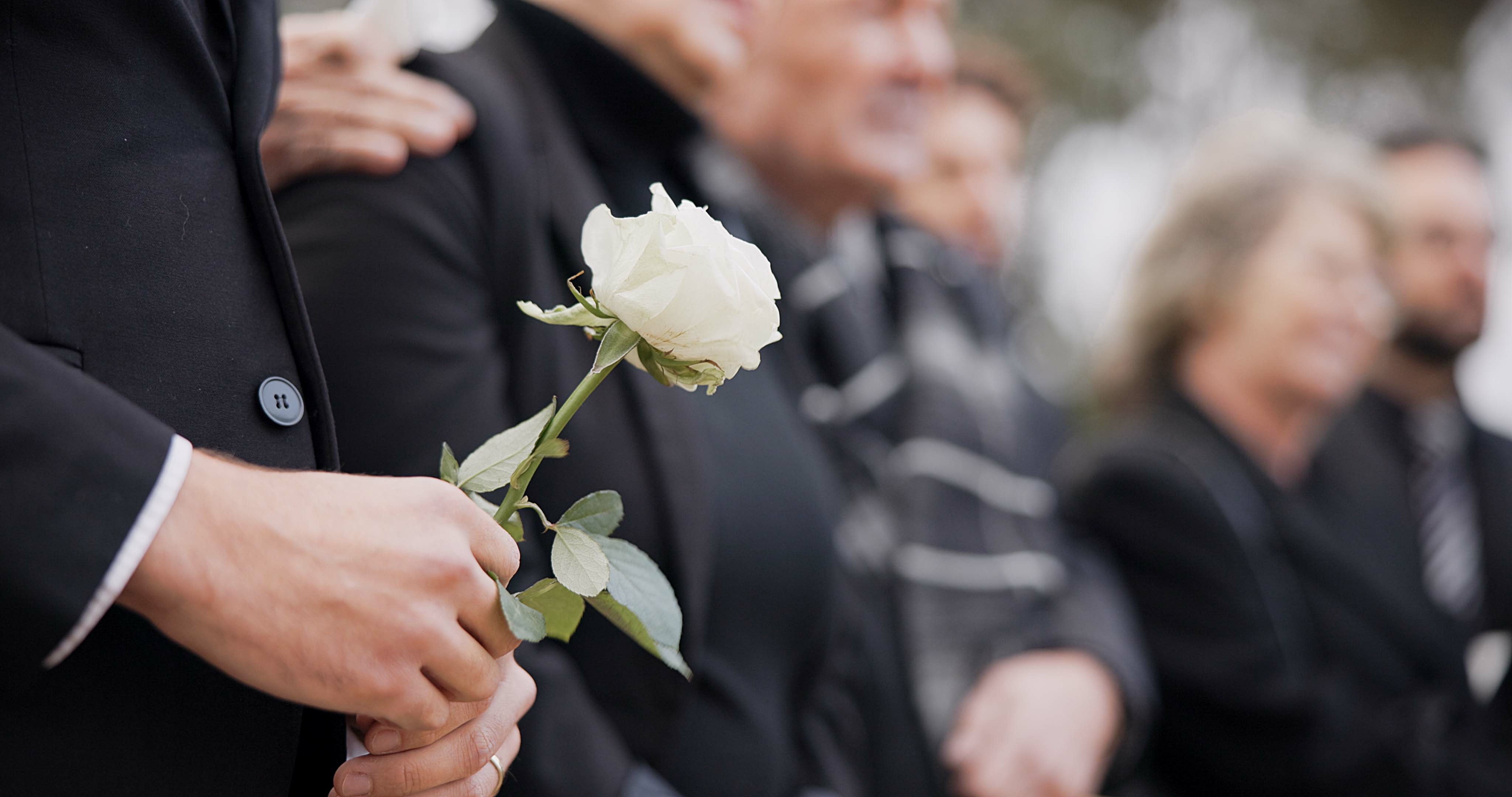 A man in a black suit holds a single white rose at a memorial as another mourner's hand rests gently on his shoulder in support